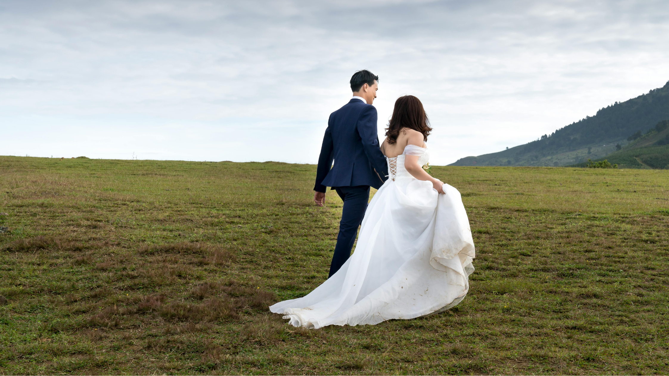Couple in wedding attire standing on a grassy field with mountains in the background